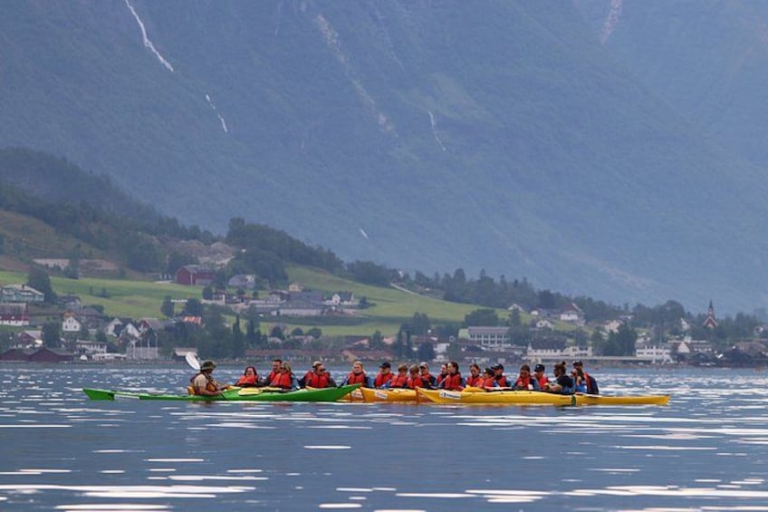 Un grupo de personas en un bote largo en un lago, con casas y montañas al fondo. 1