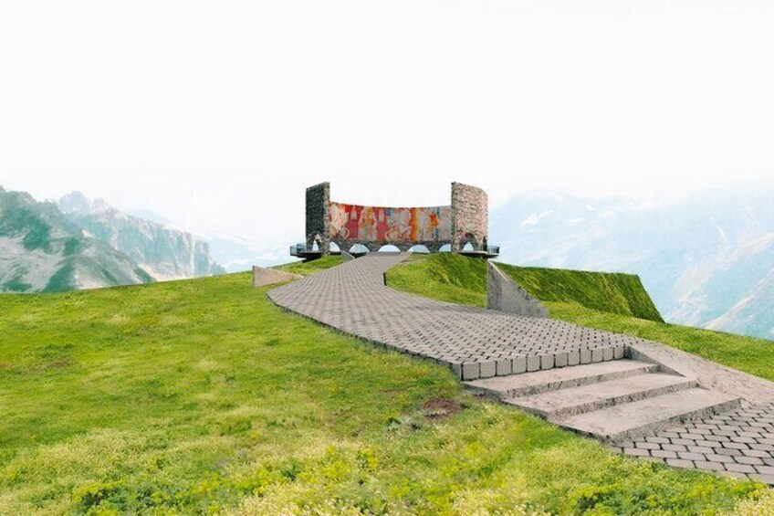 A stone pathway leading to a monument on a grassy hill with mountains in the background. 5