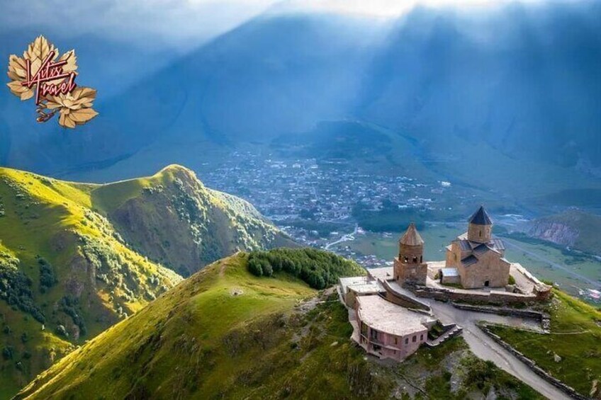 A mountainous landscape with a historic building complex, including a church with two towers, surrounded by lush green hills and a valley below. 1