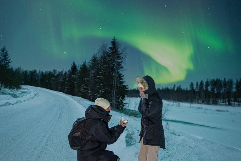 Dos personas en un paisaje nevado con carretera y árboles, bajo un cielo nocturno con auroras boreales. 1