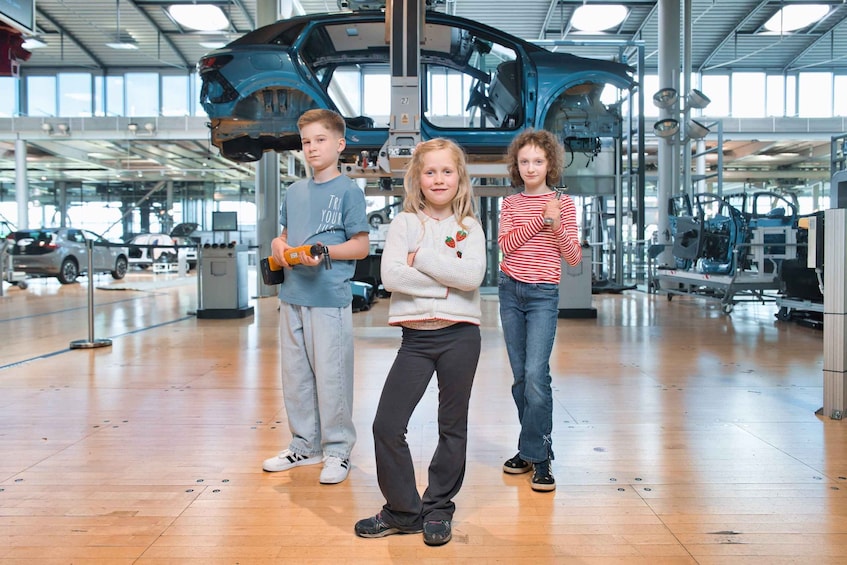 Three children standing in a car manufacturing facility with a partially assembled car in the background. 5