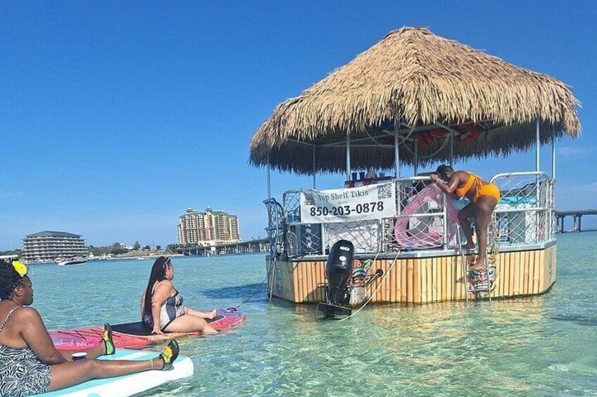 Escena de playa tropical con una estructura de techo de paja, agua azul cristalina y edificios al fondo. 3