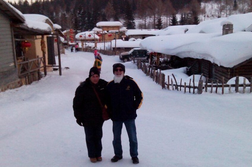 Two people standing in a snowy village with wooden houses and a fence. 4