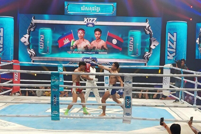 A boxing ring with two boxers in mid-fight, a KIZZ advertisement, and a crowd watching. 1