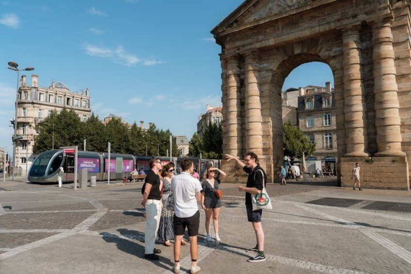 A group of people standing in front of a large stone archway with a modern tram in the background. 1