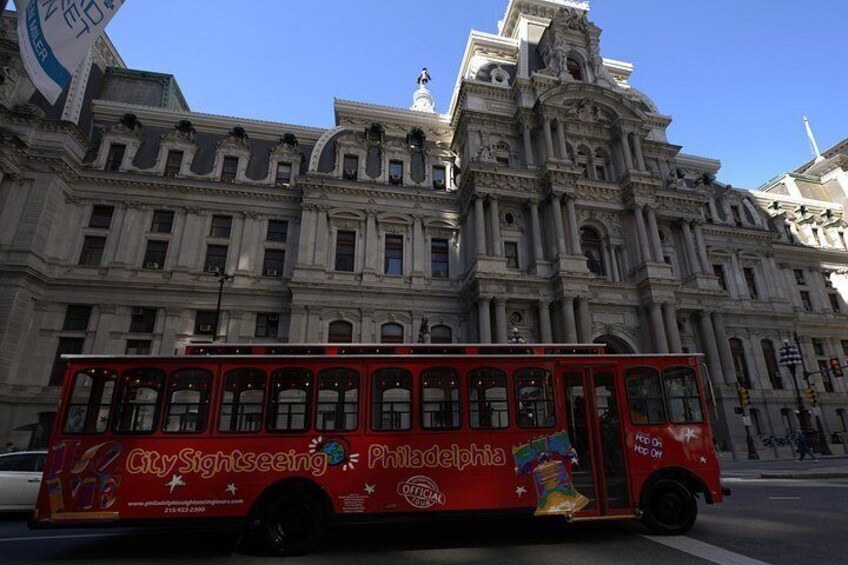 Un autobus rouge, portant l’inscription « Philadelphia City Sightseeing » sur le côté, est garé devant un grand bâtiment orné, de style architectural classique. 2