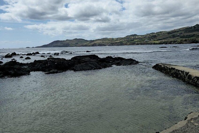 A rocky shoreline with calm water, a stone wall, and distant hills under a cloudy sky. 3
