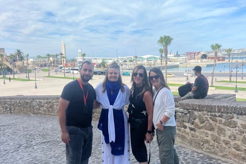 A group of people standing by a waterfront with a stone wall, palm trees, and a distant view of a cityscape. 3