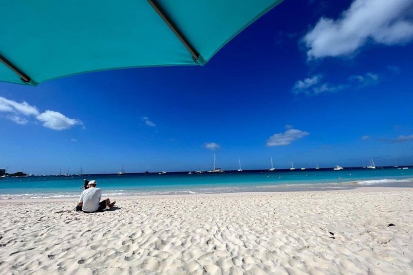 A sandy beach with a person sitting under a blue umbrella, a clear blue sky, and sailboats in the distance. 2