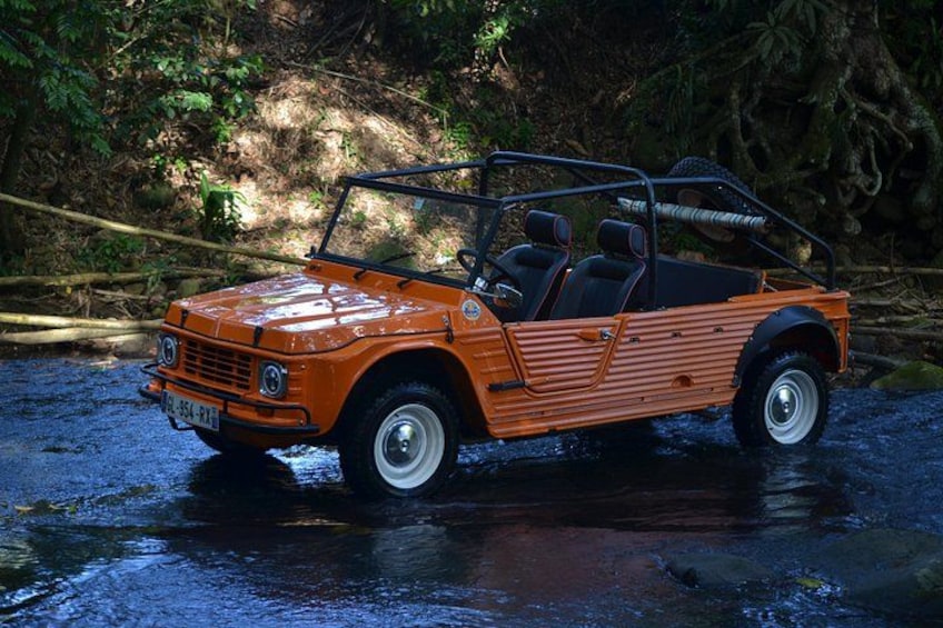 An orange vintage car with a convertible top driving through a forested area with a stream. 1