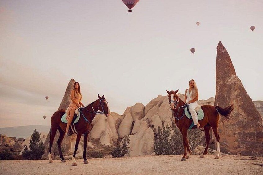 Dos personas montando a caballo en un paisaje desértico con grandes formaciones rocosas y globos aerostáticos en el cielo. 1