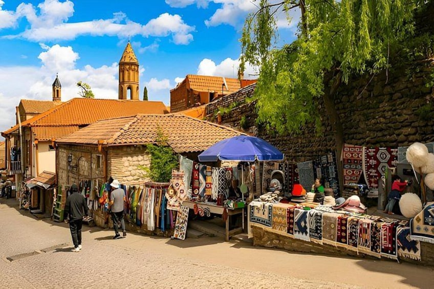 Ein Markt mit Ständen, auf denen Textilien und Kunsthandwerk angeboten werden, eine Kirche mit Kirchturm und eine Steinmauer. 3