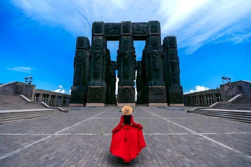 A person in a red dress and straw hat walking towards a large, dark-colored monument with intricate carvings. 1
