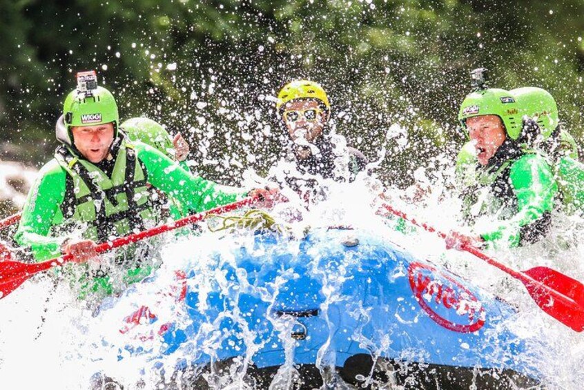 Three people in a blue kayak with red paddles, wearing green helmets and life vests, splashing water. 1
