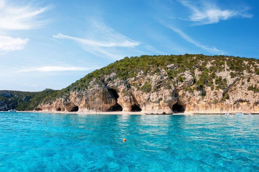 Een kustlandschap met helder turquoise water, een rotsachtige klif met grotten, en een klein bootje in de verte. 3