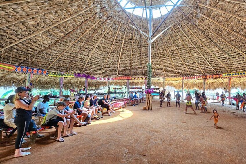 Un pavillon à toit de chaume où des personnes sont assises et debout, entouré d’une zone sablonneuse. 2