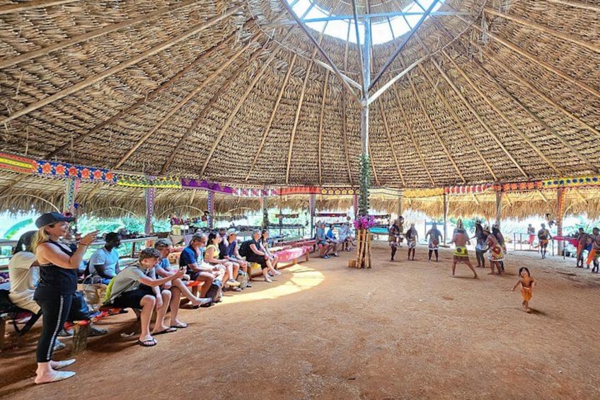 A thatched-roof pavilion with people seated and standing, surrounded by a sandy area. 2