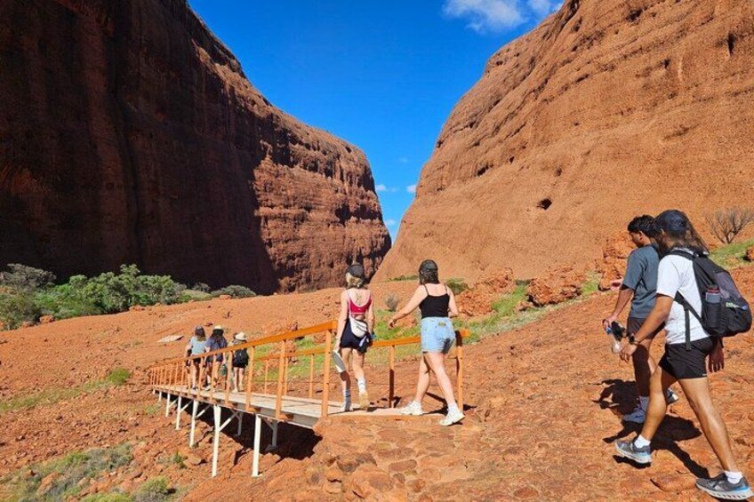 Un grupo de excursionistas en un sendero pedregoso con un puente de madera, rodeado de formaciones rocosas rojas. 4