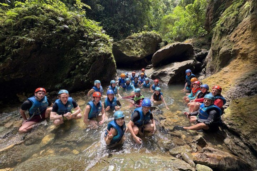 A group of people in blue life jackets sitting in a shallow stream surrounded by rocky terrain. 3
