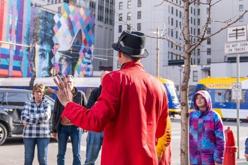 A person in a red coat and black hat gesturing, with a colorful mural and a Metro bus in the background. 3