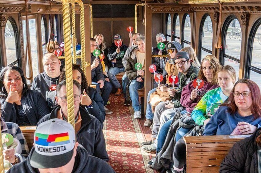 A group of people seated inside a vintage tram with ornate wooden interiors and large arched windows. 2