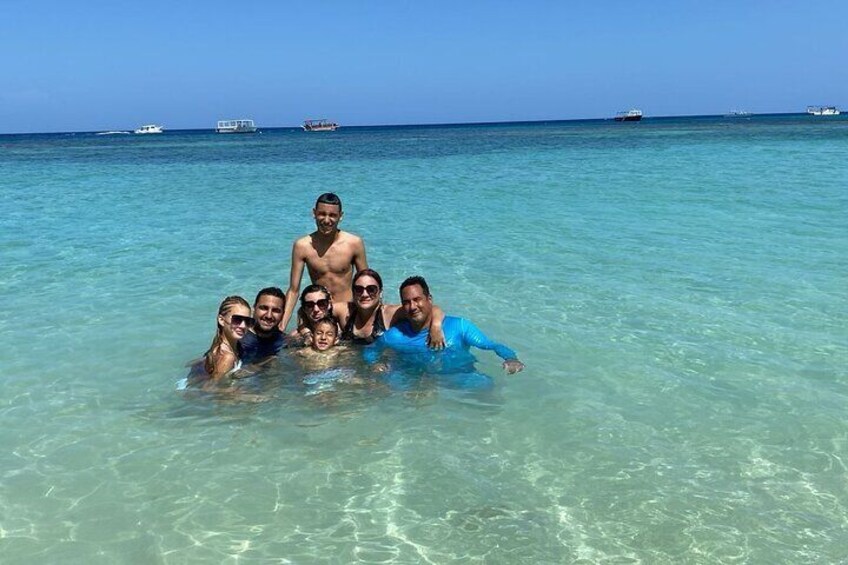 A group of people in clear turquoise water with boats in the distance. 2