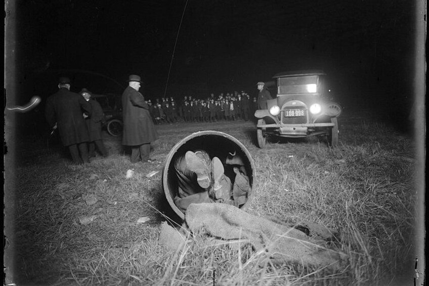 Un'auto d'epoca con i fari accesi, un gruppo di persone sullo sfondo e un grosso oggetto cilindrico a terra. 3
