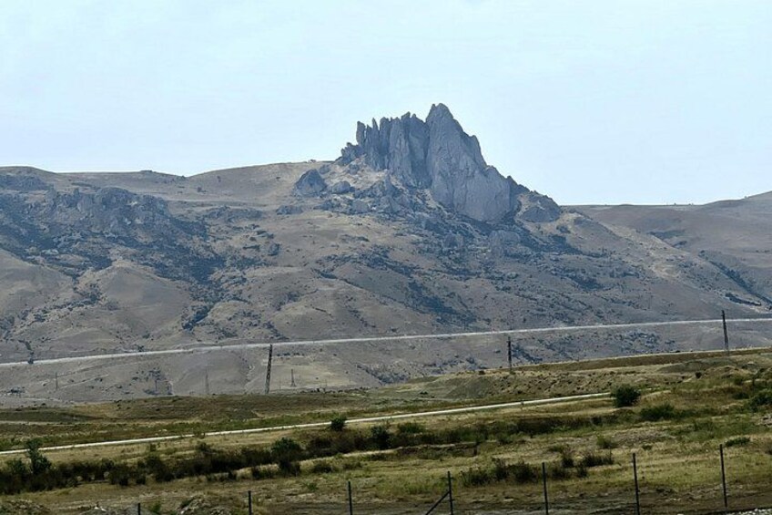 Een bergachtig landschap met een opvallende rotsachtige piek, een onverharde weg en schaarse begroeiing. 3