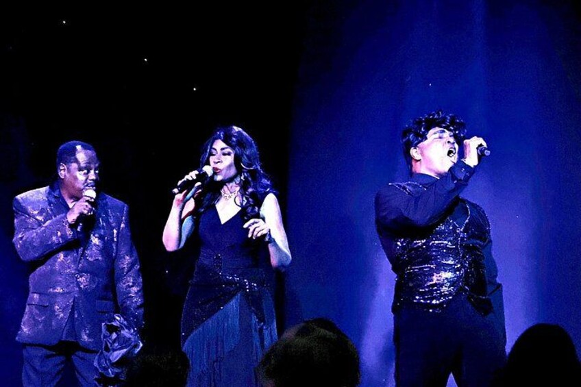 Tres personas en el escenario con micrófonos, vestidas con atuendos oscuros y brillantes. 2