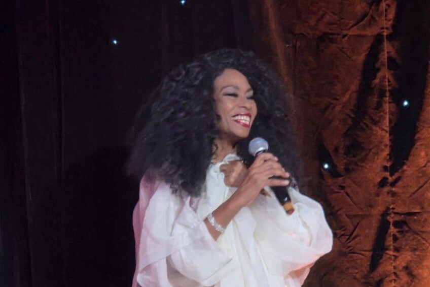 A person holding a microphone, dressed in a white outfit, with a dark background and a tree-like structure to the right. 3