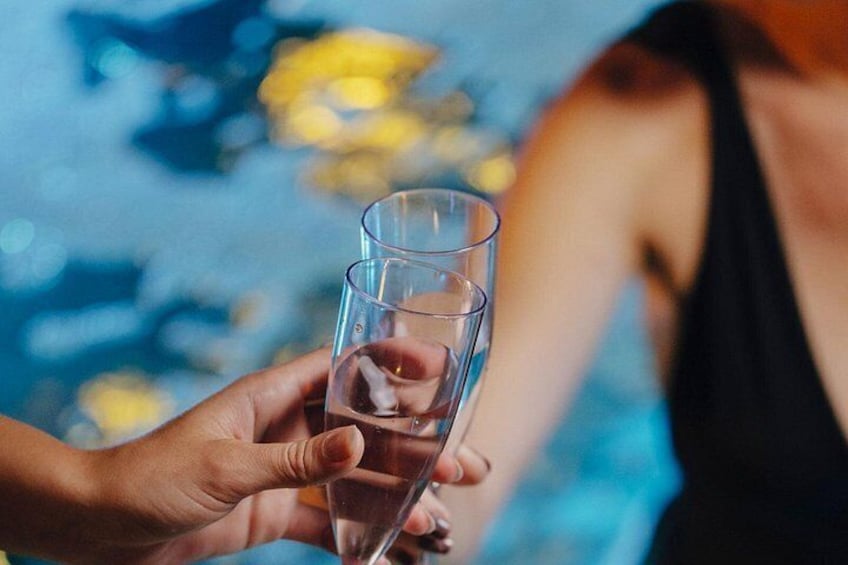 Une main tenant un verre transparent contenant un liquide, possiblement du champagne, près d'une piscine, avec de l'eau et des fleurs jaunes floues en arrière-plan. 4