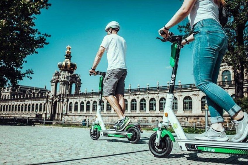 Dos personas montan patinetes eléctricos sobre una superficie pavimentada, con un edificio histórico al fondo. 3
