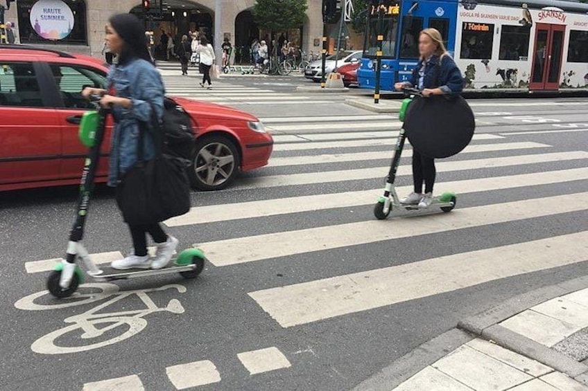 Dos personas montando scooters en un paso de cebra, con un coche rojo y un autobús al fondo. 4