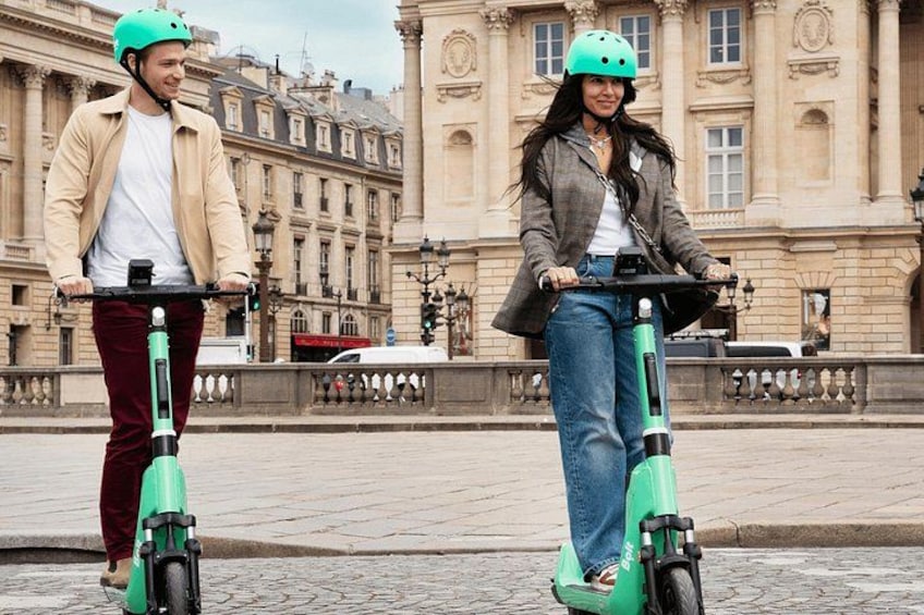 Dos personas montando scooters eléctricos sobre una superficie pavimentada, con un edificio histórico al fondo. 1