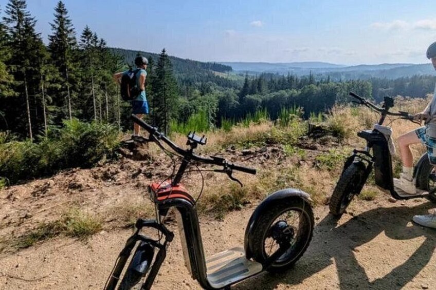 A dirt path with two electric scooters, a person standing, and a forested landscape in the background. 4