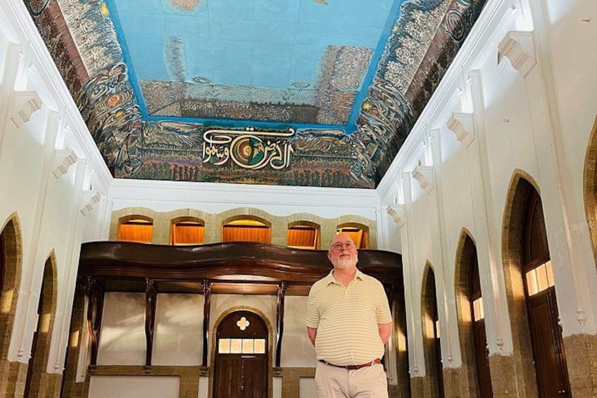 A man standing in a hallway with a ceiling mural, arched windows, and a wooden door. 5