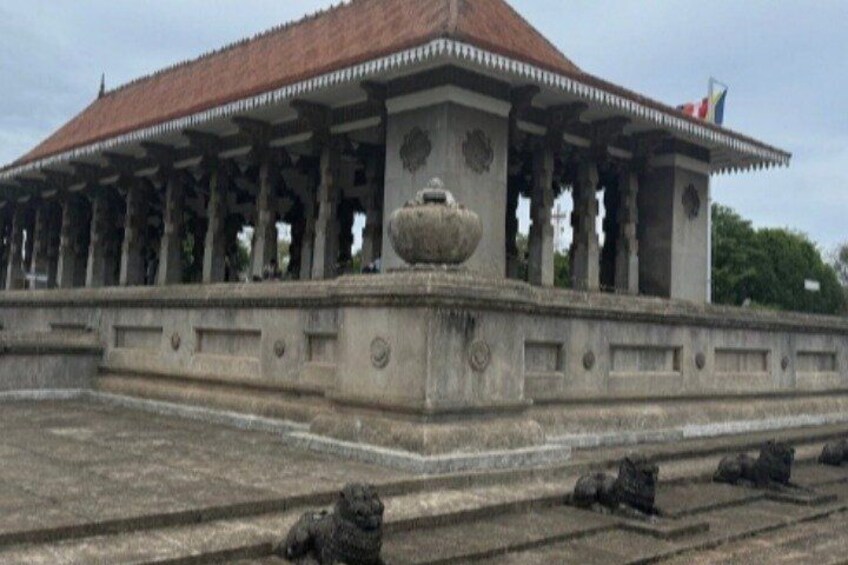 Eine große, offene Halle mit einem Ziegeldach und Säulen, umgeben von einer niedrigen Mauer und einem großen Steinkrug. 5