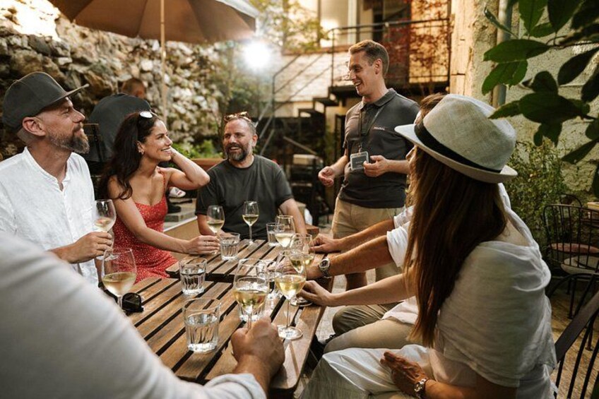 A group of people sitting around a wooden table outdoors, holding wine glasses. 2
