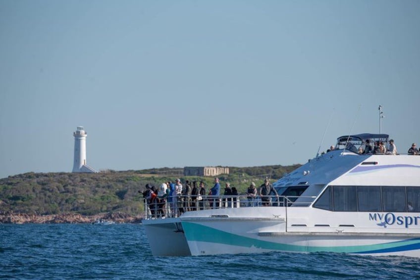 A boat with people on deck, a lighthouse in the background, and a building on the shore. 1