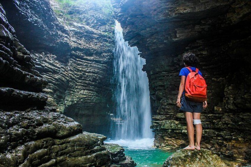 A person standing on a rock with a red backpack, looking at a waterfall in a rocky gorge. 4