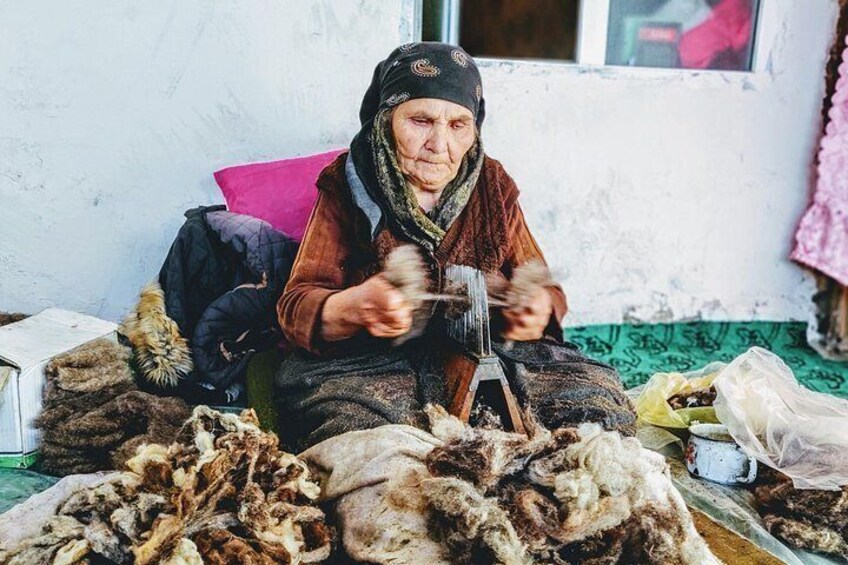 An elderly person seated on a chair, engaged in a manual task with a tool, surrounded by a cluttered environment with various items and a window in the background. 3