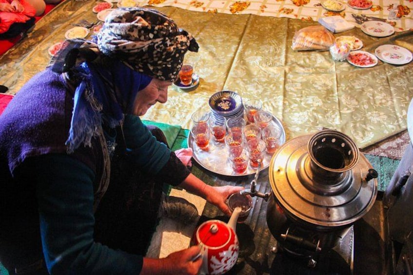 A person preparing tea with a samovar and teapot, surrounded by various food items and utensils on a table. 1