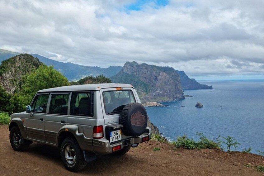 A silver SUV parked on a dirt path overlooking a coastal area with mountains and the ocean. 2