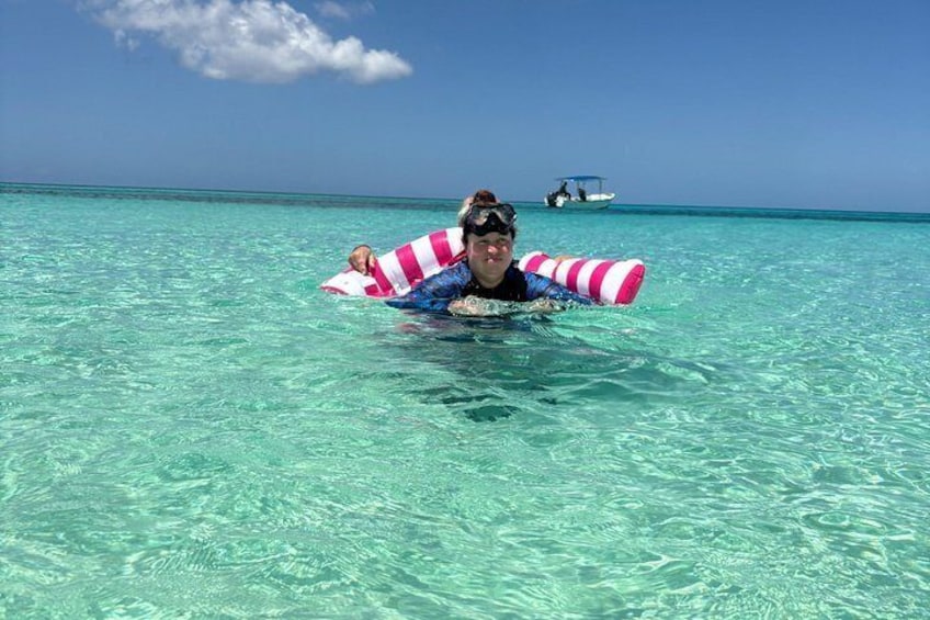 Une personne flotte sur une plage, allongée sur un flotteur rayé de rose et de blanc, dans une eau turquoise cristalline, et on aperçoit un bateau au loin. 4