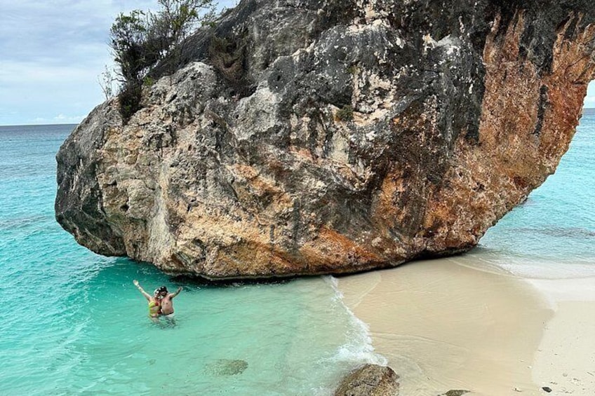 Une personne nage près d’une grande formation rocheuse sur une plage où l’eau est d’un bleu turquoise limpide. 3