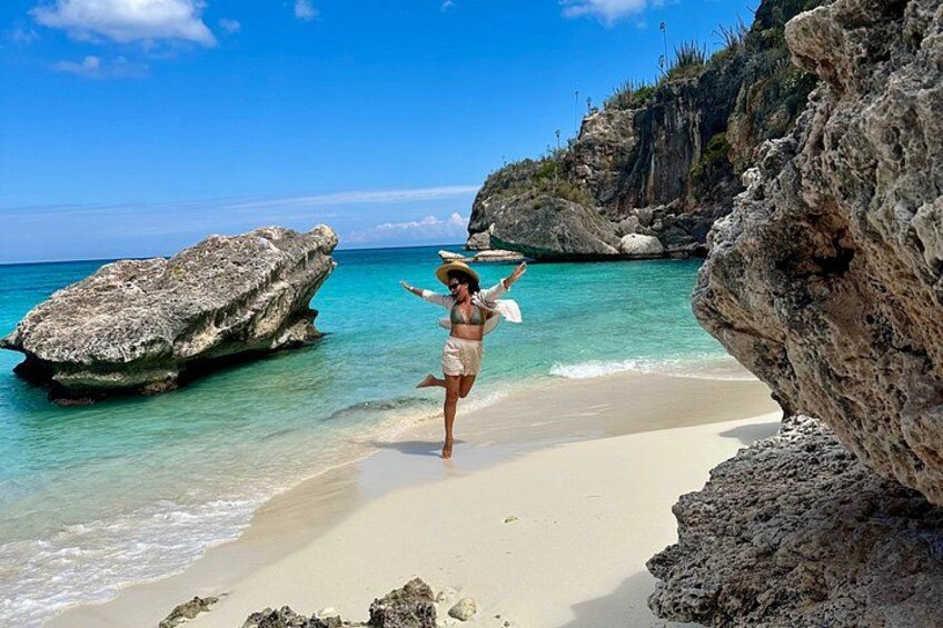 Une personne en bikini sur une plage de sable fin, avec une eau turquoise et limpide, de gros rochers et une falaise rocheuse en arrière-plan. 1