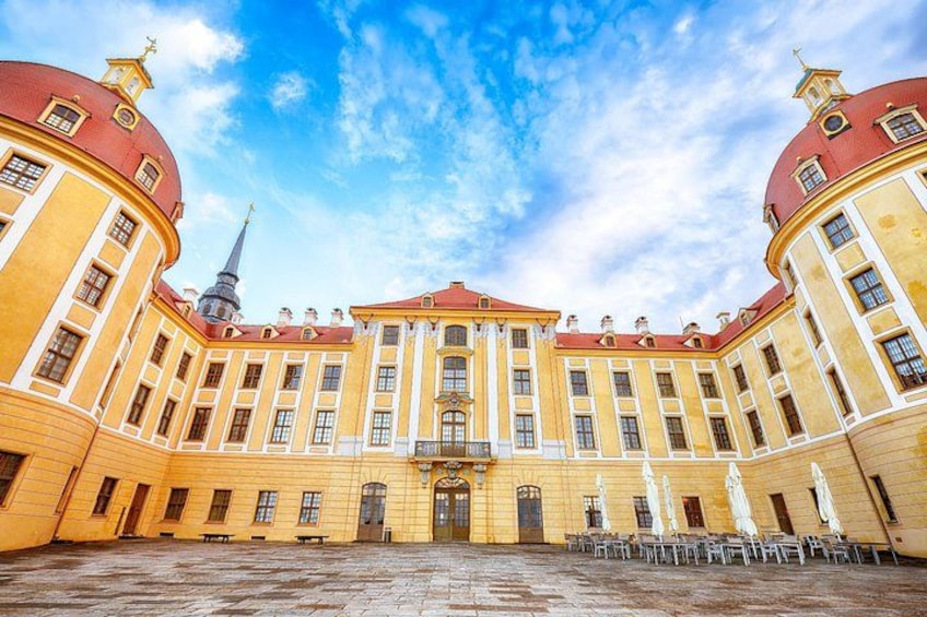 A large yellow building with a central entrance, surrounded by a courtyard with tables and umbrellas. 2