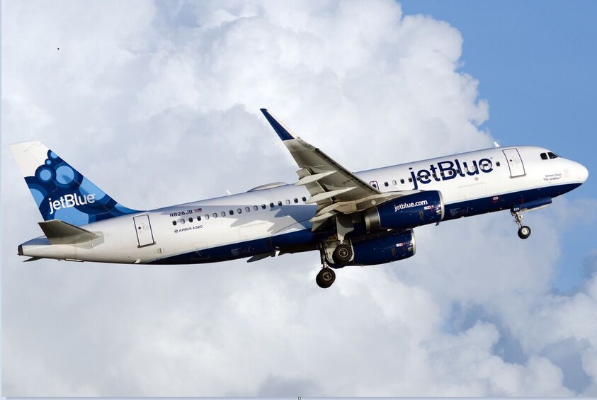 Sinivalkoiseksi maalattu JetBlue-lentokone lentää, taustalla näkyy pilviä ja kirkas taivas. 4