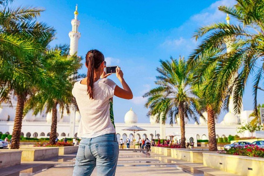 A person taking a photo with a camera, palm trees, and a white building with a dome in the background. 1