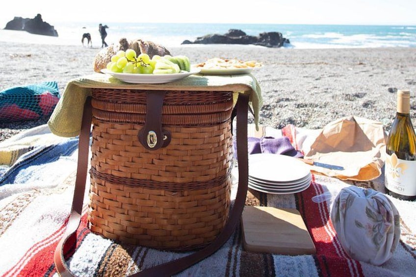 A picnic setup on a beach with a basket, plates, a bottle, and a blanket. 1